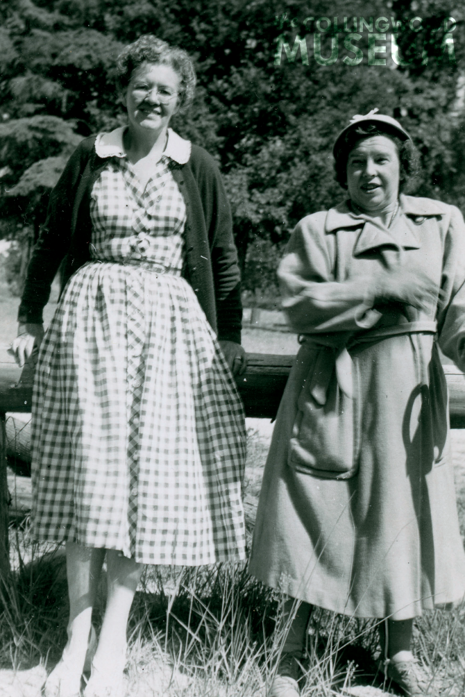 Two women stand together in front of evergreen trees 