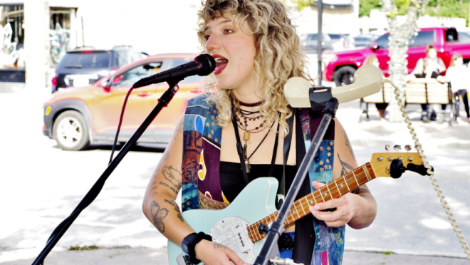 A performer with curly blonde hair sings into a microphone while playing a light blue electric guitar. They wear a colorful patchwork vest over a black top and have tattoos on their arms.