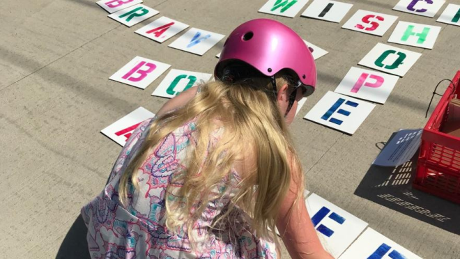 A child wearing a pink helmet is arranging letter stencils on the ground, spelling out "HOPE" and "BRAVE".