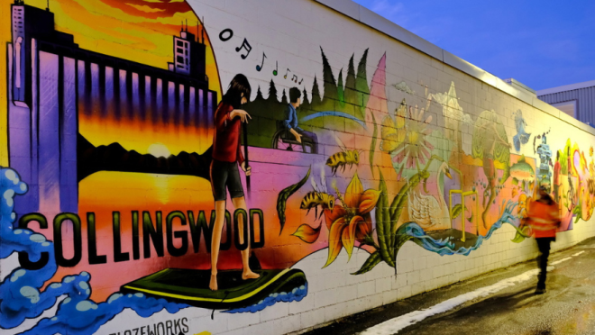 A vibrant mural on a brick wall depicts the word "COLLINGWOOD" in large letters, with a person on a surfboard in front of a sunset, and various colorful illustrations of nature and urban elements, including bees, flowers, and buildings.