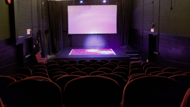 A view from the audience seats looking towards a small stage with a projection screen and a patterned rug on the floor. The stage is lit with purple lights.