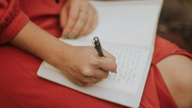 A person wearing a red dress is writing in a notebook with a pen.