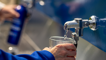 A person fills a glass with water from a drinking fountain, with a blue water bottle in the background.