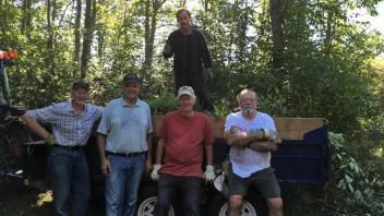 A group of five individuals stand in front of a trailer filled with plants and debris, with trees in the background.