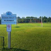 A sign for "Fisher Field #3" stands in front of a soccer field with a goal net and tree line the background.