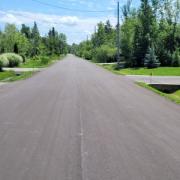 A long asphalt road that stretches into the distance, lined with green trees and lawns under a bright blue sky.