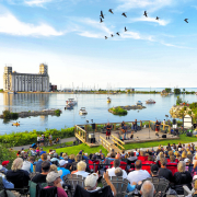 An outdoor concert is taking place in front of a large body of water with a grain elevator in the background. Many people are seated on the grass watching the performance.