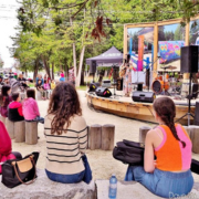 An outdoor concert stage with performers and a seated audience in the foreground.