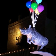 sculpture of a rhinocerous with a bundle of brightly coloured balloons attached to its midsection under a night sky