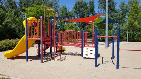Georgian Meadows Park Playground Equipment Red playground structure with a yellow slide and climbing system. Three shade sails in background. Sand surface.