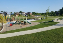 View of entire park from grass hill showing three playground structures, pavilion and neighborhood in the background.