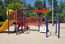 Red playground structure with a yellow slide and climbing system. Three shade sails in background. Sand surface.