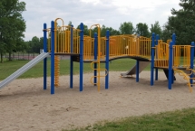 Blue and yellow playground with two slides. There is a set of climbing equipment. Playground surface is sand.