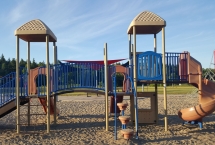 Brown and blue playground equipment with three slides and climbing equipment. set of swings and climber in the background. Playground has sand surface.