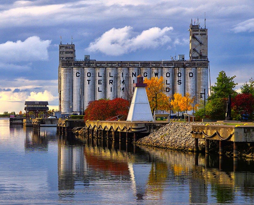 The Collingwood Terminals Limited grain elevators by the waterfront, surrounded by vibrant autumn trees and a small white lighthouse under a cloudy sky