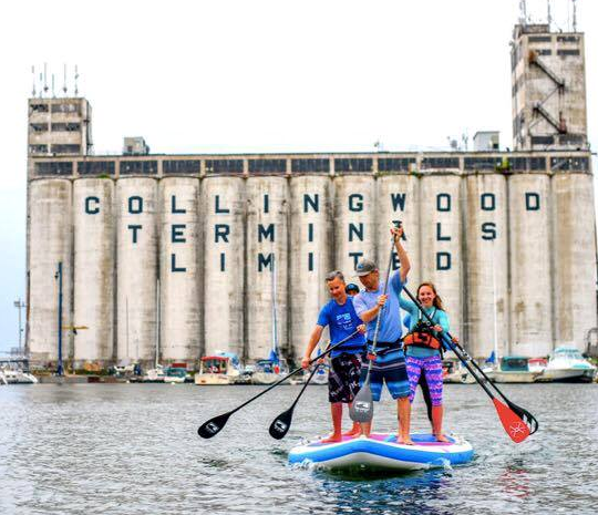 People paddling on a large stand up paddleboard with the Terminals building in the background